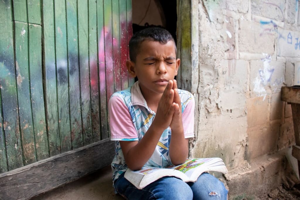 Luis is wearing a blue and white patterned shirt with pink sleeves and jeans. He is sitting outside his home praying with his hands pressed together in front of him. On his lap is his Bible.