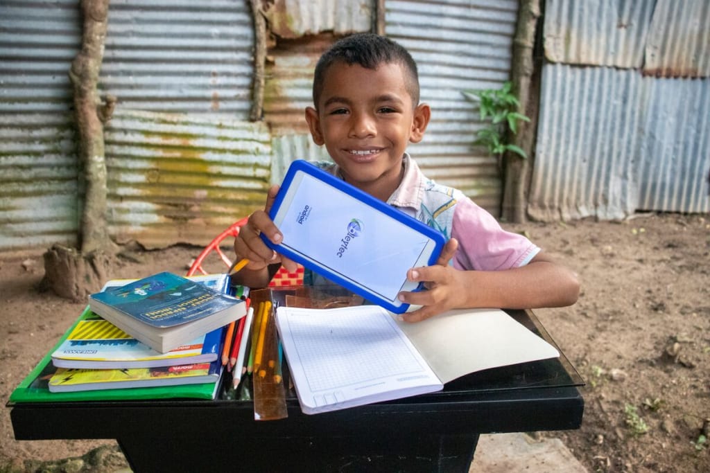 Luis is wearing a blue and white patterned shirt with pink sleeves and jeans. He is sitting at a desk outside his home and is holding a tablet he uses for virtual classes. On the desk in front of him are books, notebooks, and pencils.
