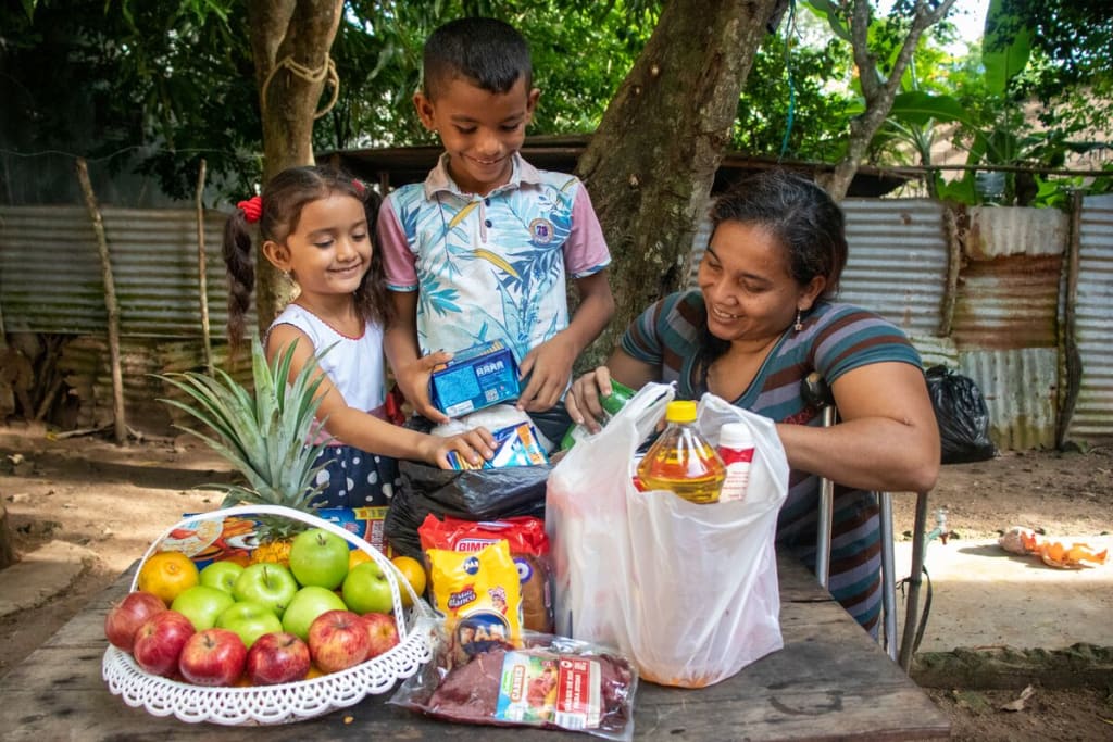Luis is wearing a blue and white patterned shirt with pink sleeves and jeans. He is in his back yard with his mother and sister. They are looking through food delivered to them by Compassion.