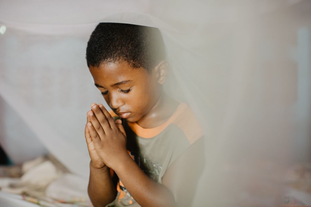 A boy in a grey shirt folds his hands in prayer under a mosquito net