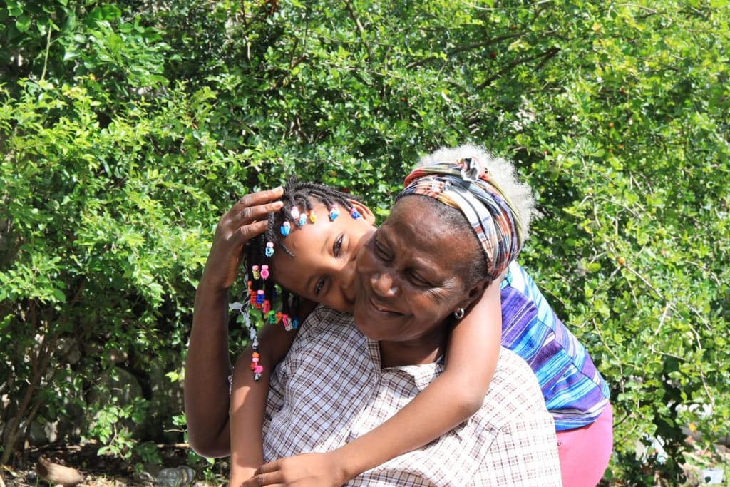 A young girl stands behind her grandmother and kisses her cheek