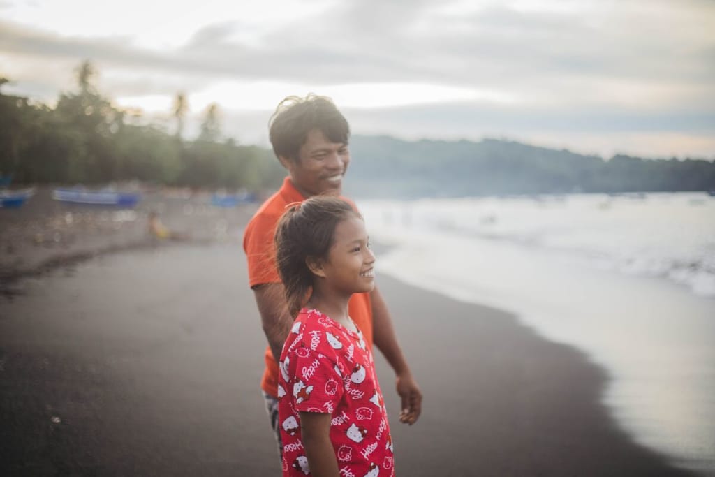 Keilah is wearing a red and white shirt. She is walking along the beach with her father, Juandris, wearing an orange shirt.