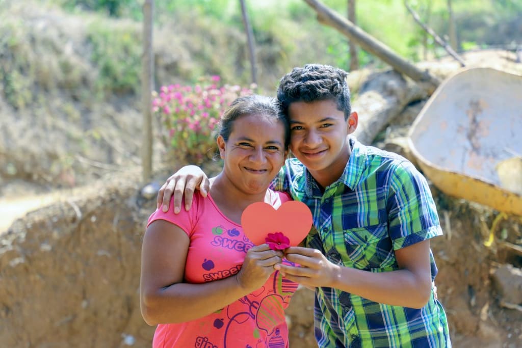 A boy in a blue and green shirt and his mother in a pink shirt hold a heart shape and a flower as they smile.