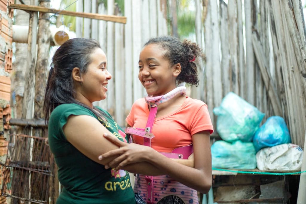 A girl in an orange shirt hugs her mother in a green shirt. They look at each other and smile.