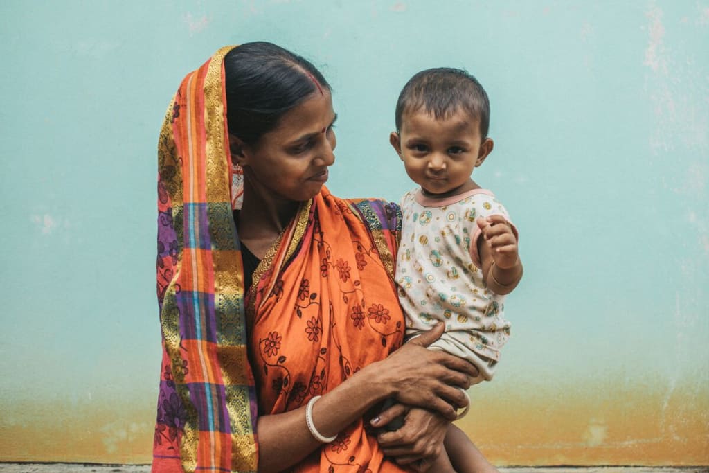 A mother is standing in front of a light blue wall wearing an orange saari with her child in her arms. She is looking at her child and smiling.