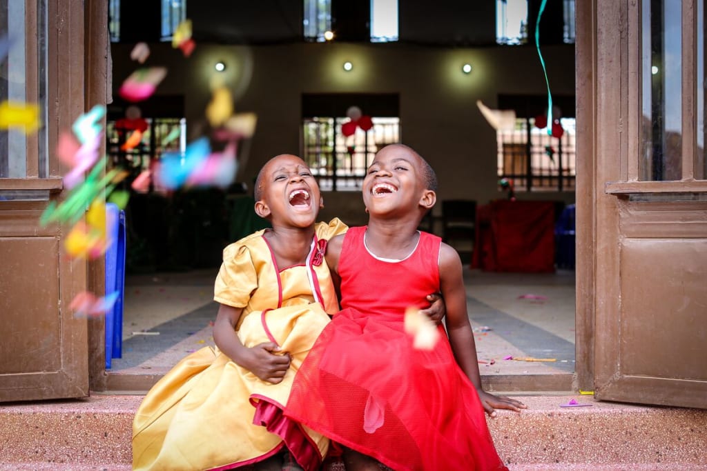 Two girls are seen here, seated in an open doorway to the church/project. The girls are both wearing dresses, one is wearing yellow and the other is wearing red. They are smiling and have their arms around one another, and have tossed confetti into the air.