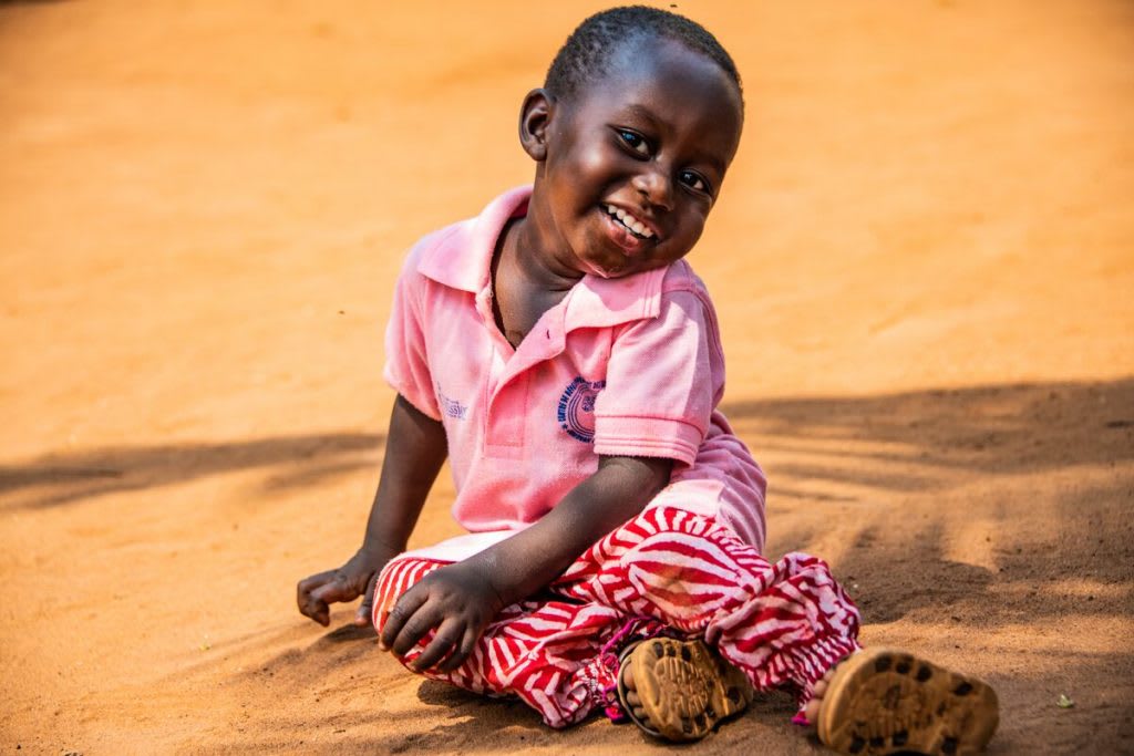 A little boy sits on the sandy ground smiling. He is wearing a pink polo shirt and red patterned pants.