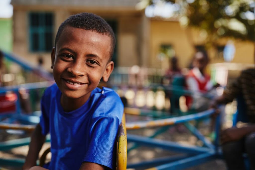 A young boy wears a blue t-shirt and sits on playground equipment smiling.