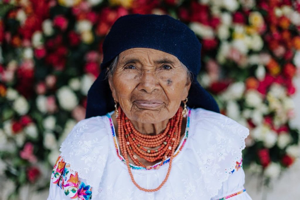 A elderly woman is wearing a necklace of strings of orange beads. She has a white shirt with flowers and has a blurred background of flowers behind her.