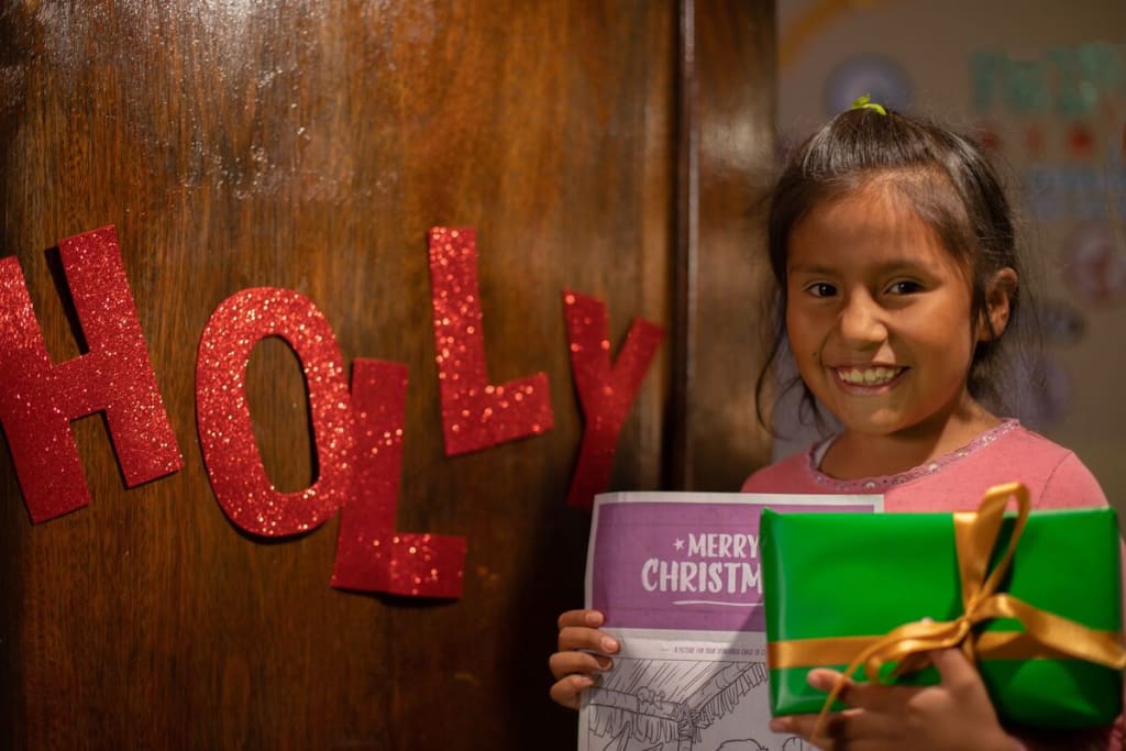 A girl wearing pink stands with a letter and a gift in front of the word "holly"