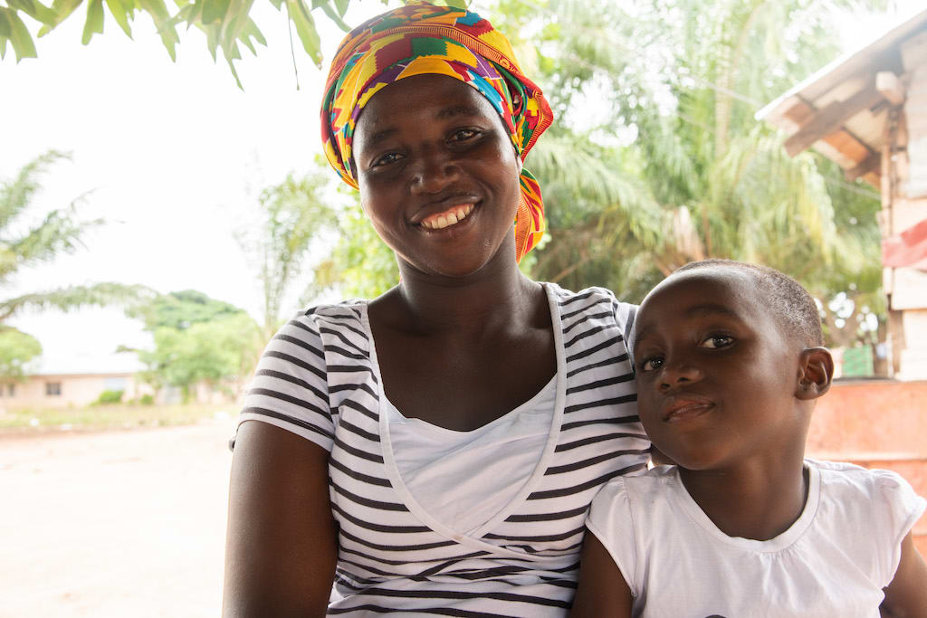 In Ghana, 5-year-old Emmanuella, wearing a white shirt, is sitting outside her home with her mother, Diamatu, wearing a black and white striped shirt and a colorfully patterned head covering.