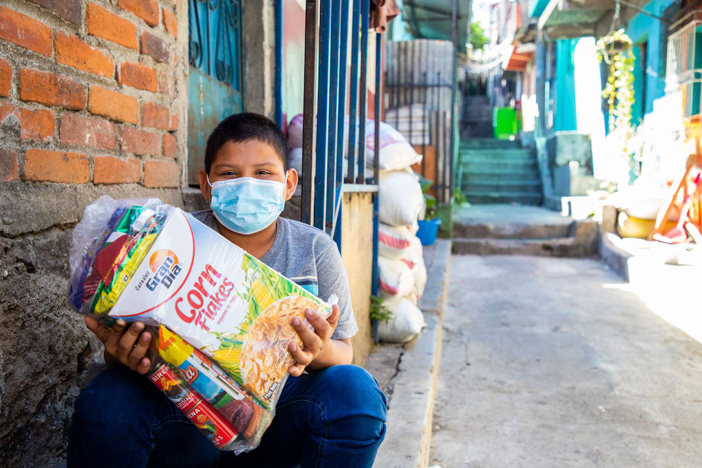 In El Salvador, 11-year-old Jhonny is sitting on the steps in front of his home and is holding a bag of food he was given by the Compassion centre. He is wearing a face mask.