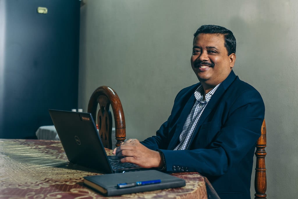 Michael is wearing a blue jacket and is sitting at his table working on his computer. He is working from home.