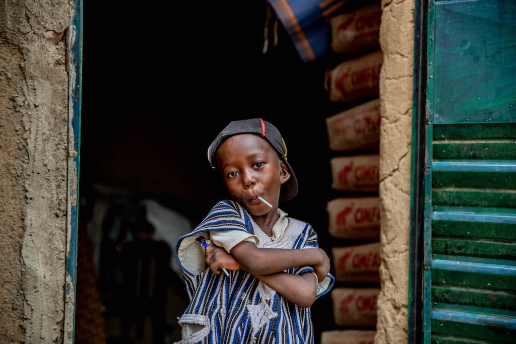 Four-year-old Achille is wearing a gray, black, and navy striped shirt and blue pants. He is standing in the doorway of his home and has a sucker in his mouth.