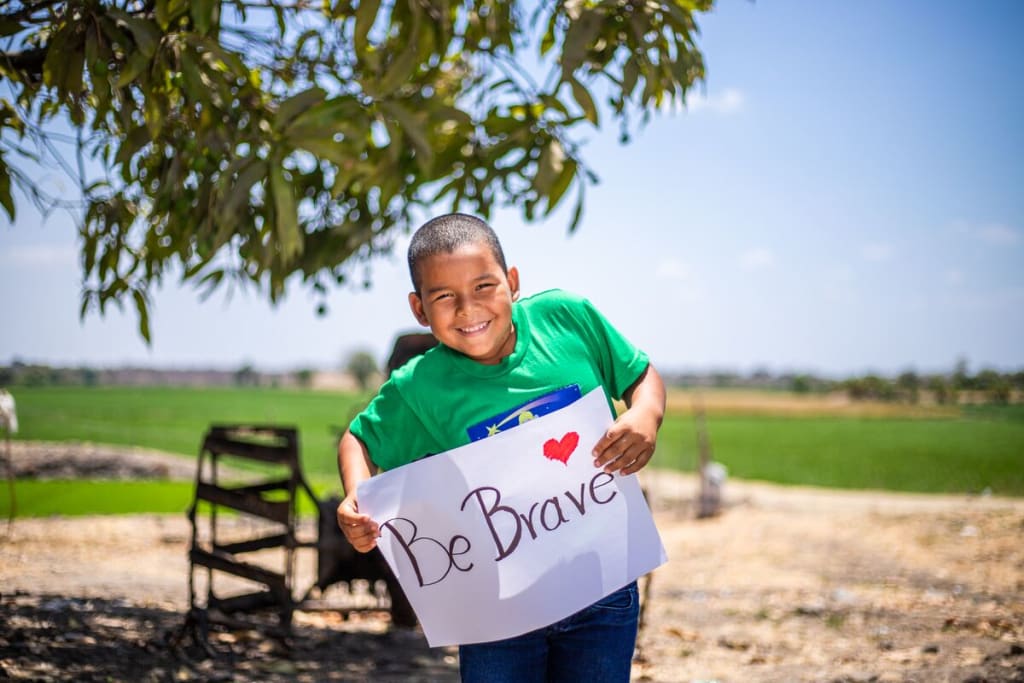 Little boy wearing a green shirt holds a sign that says, "Be Brave"