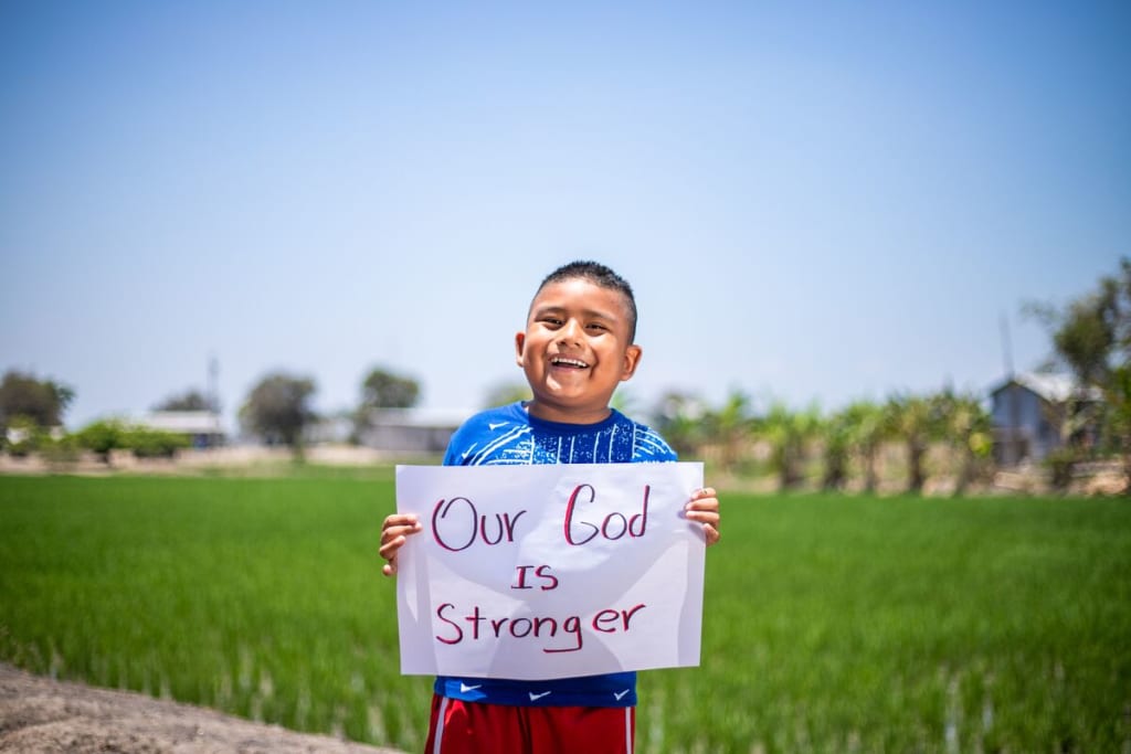 Little boy wearing a red shirt holds a sign that says, "Our God is stronger."