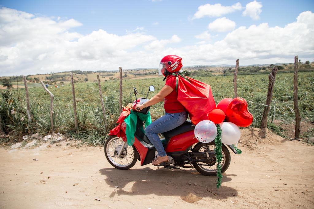 A volunteer is on a motorcycle wearing a red cape, a red helmet, with balloons and streamers flying off of it.