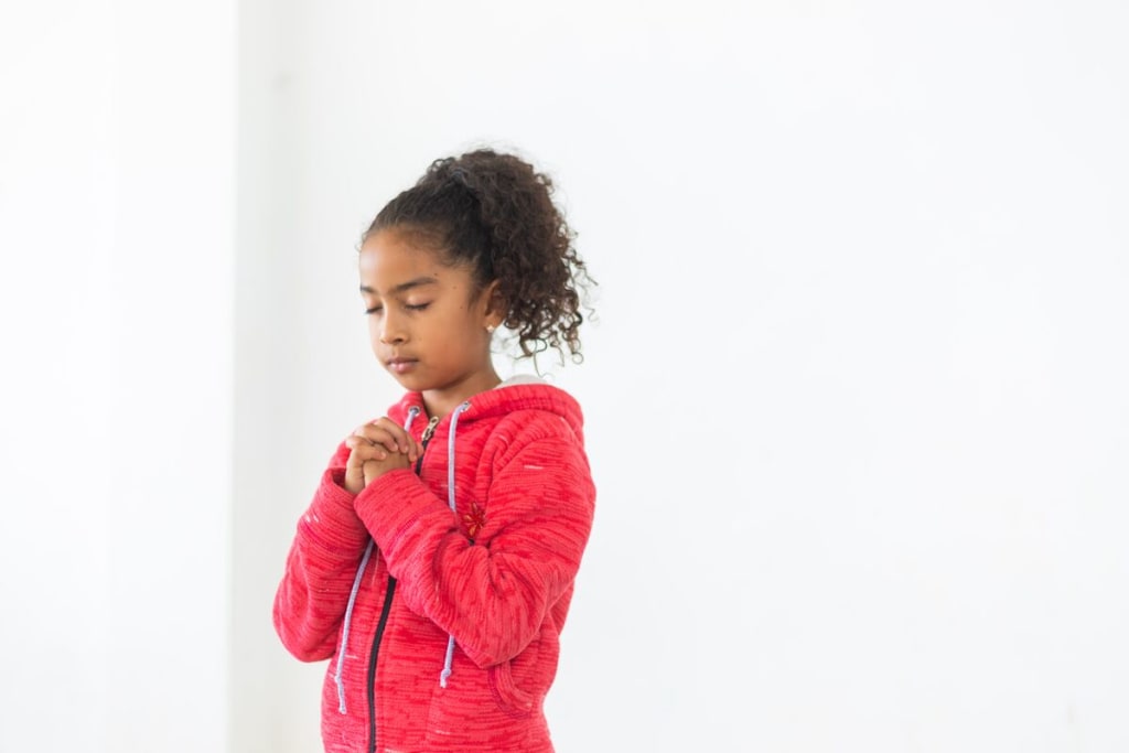 A girl in a pink sweater prays in front of a white wall