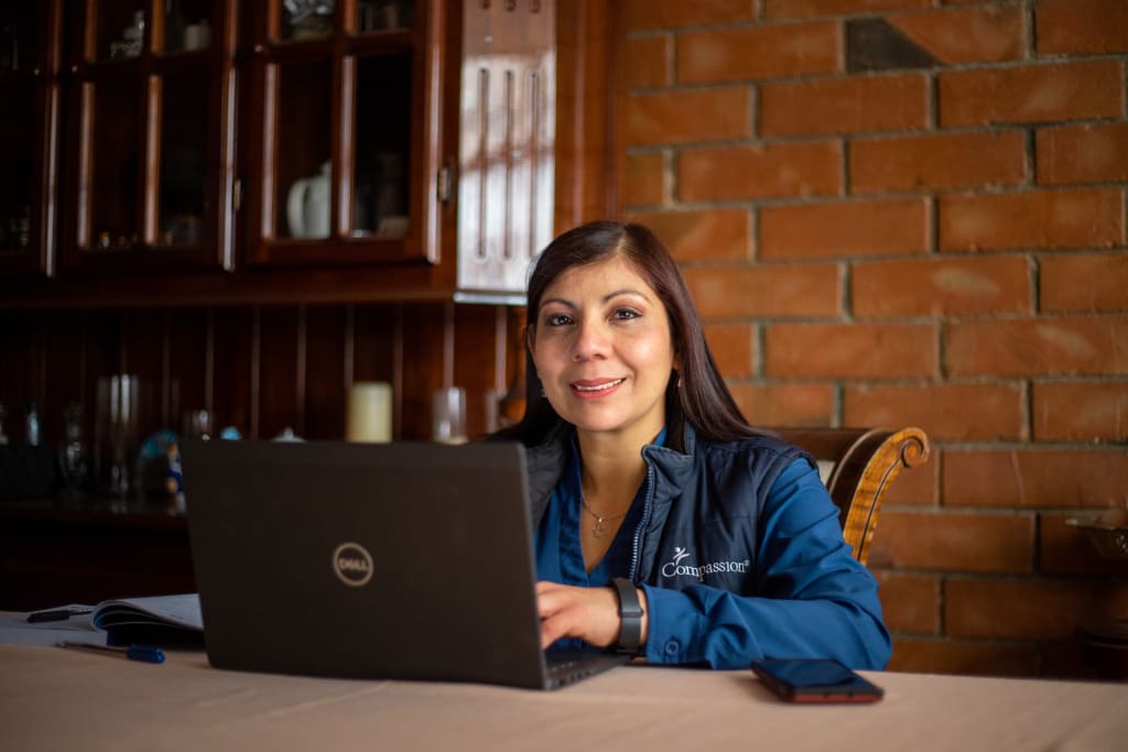 Project Facilitator Virigina sits at her desk behind a Dell laptop. She is wearing a blue jacket with the Compassion logo on it.