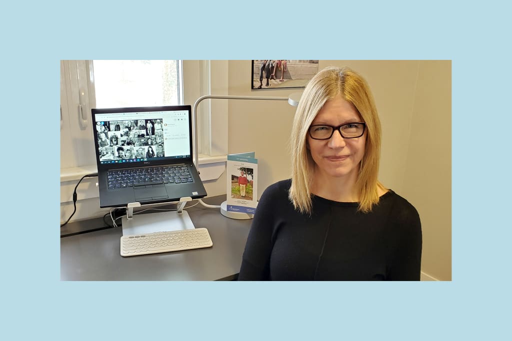 Teresa is wearing glasses and a black shirt, sitting at her desk with her laptop and a photo of her sponsored child behind her.
