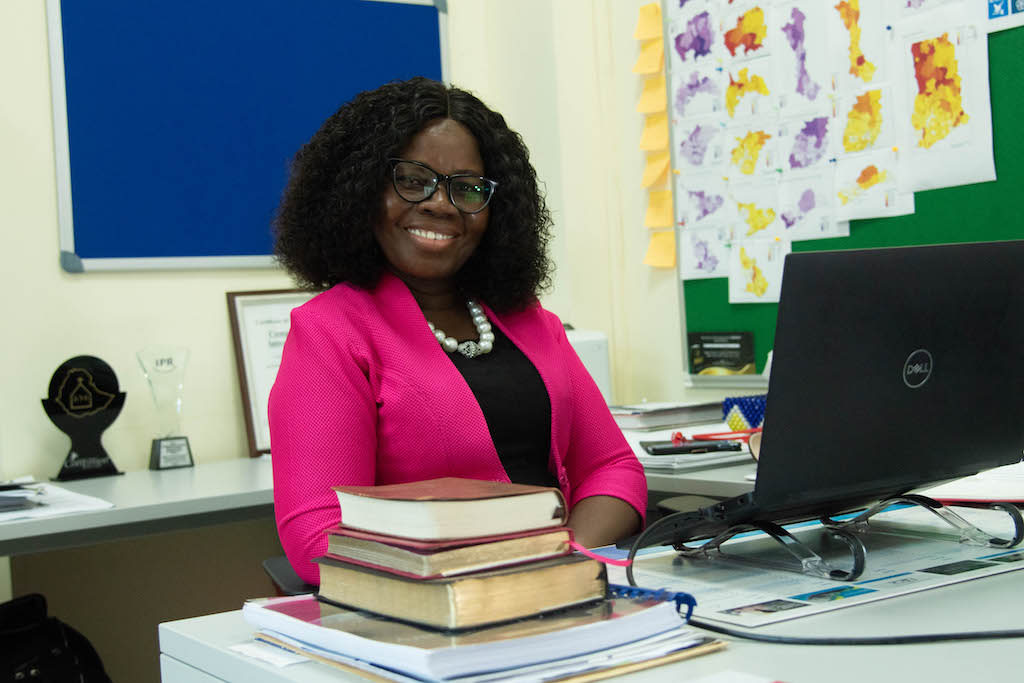 National Director for Ghana, Gifty Appiah, is wearing a black dress and a bright pink jacket. She is sitting at a desk in front of a computer.