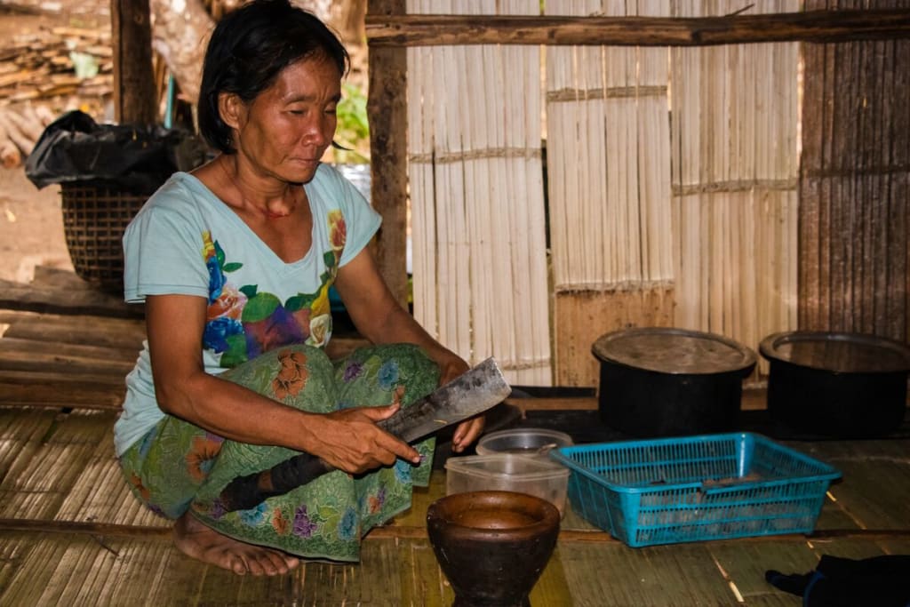 A woman wearing a blue shirt crouches in her home with a knife cutting food to eat. She's in a bamboo home.