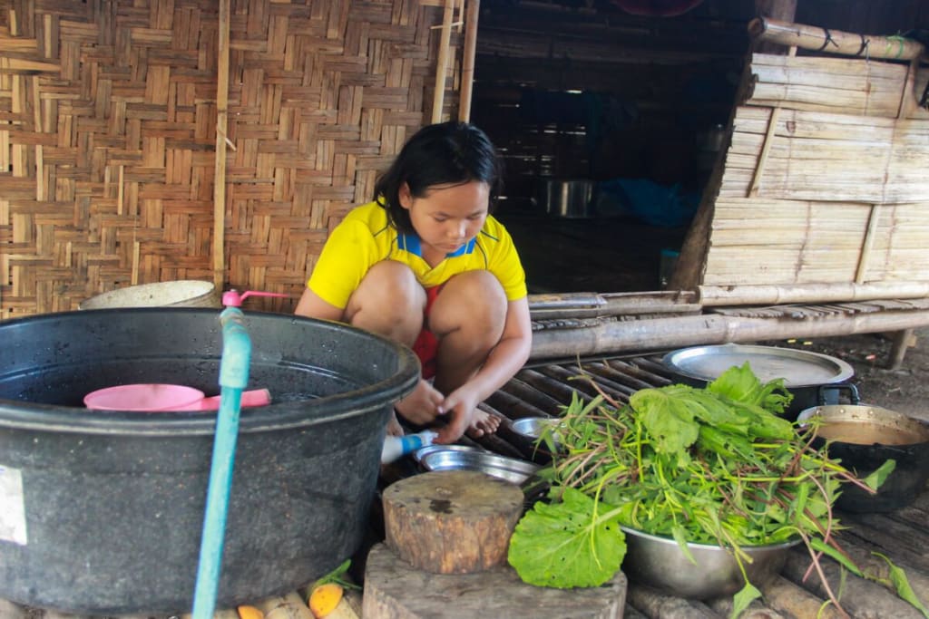 Young girl in a yellow shirt crouches to wash vegetables outside.