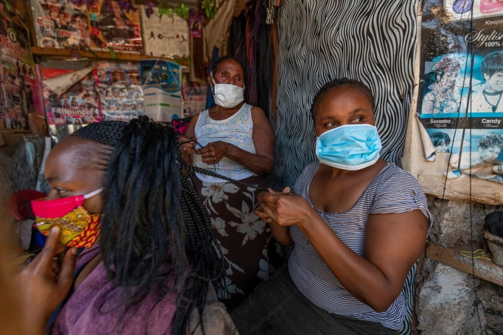 Two women wearing masks braid another woman's hair at a home salon