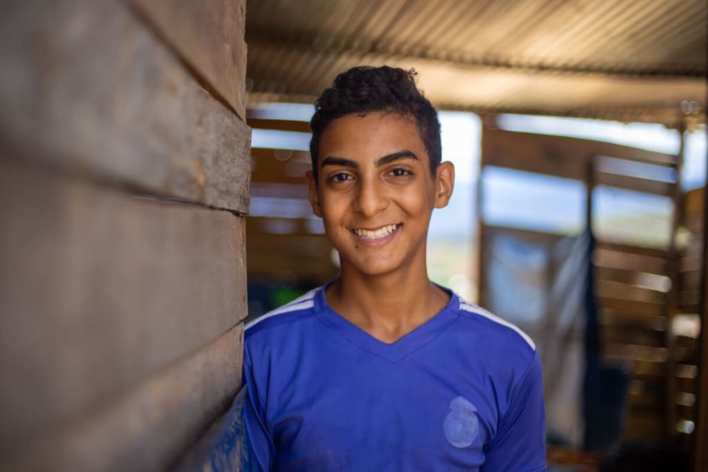 A teenage boy stands in a wooden shack. He smiles, happily.
