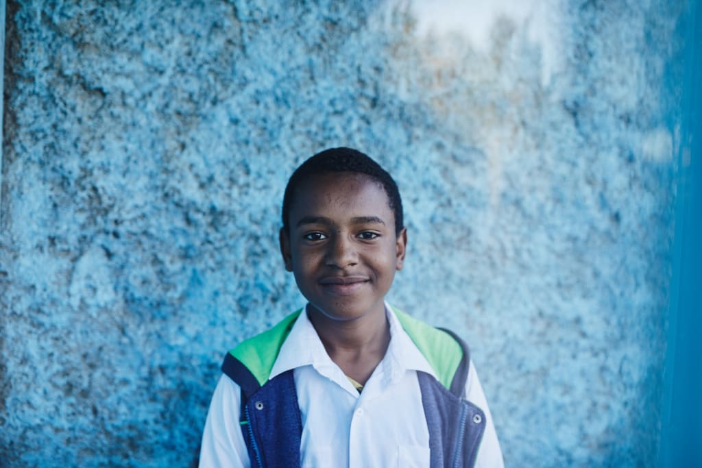 A portrait of an Ethiopian teen, against a blue wall wearing a white shirt and blue vest.