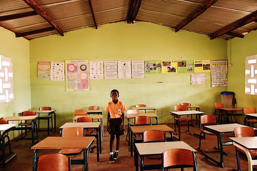 A boy stands amongst desks and chairs in a classroom.