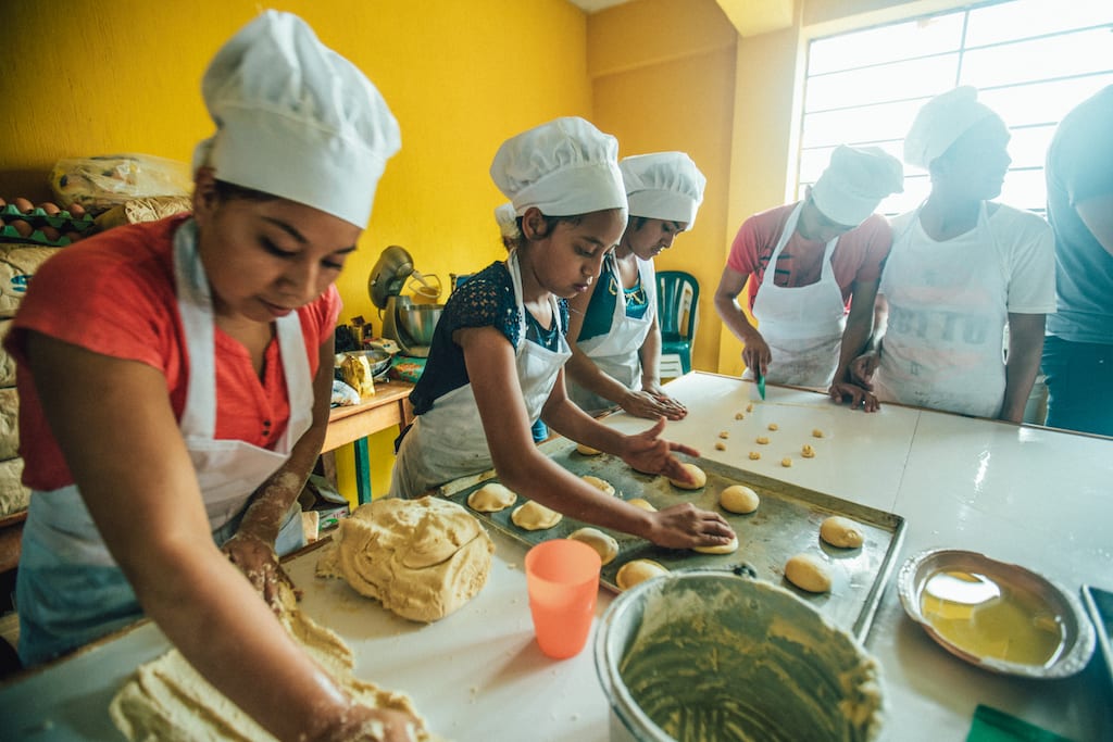 A group of students wearing aprons and chef's hats roll out dough.