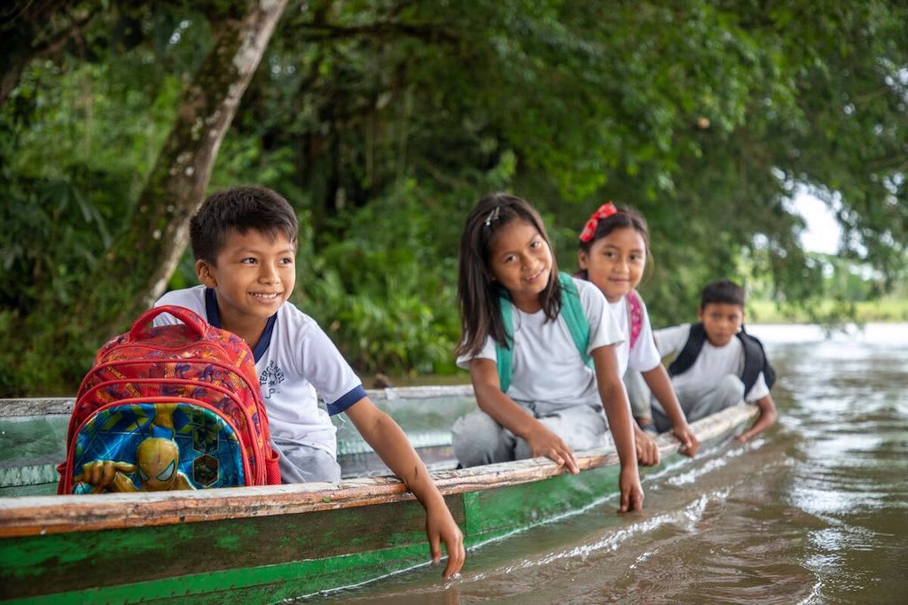 A group of four children sit in a canoe with their backpacks, on their way to school.