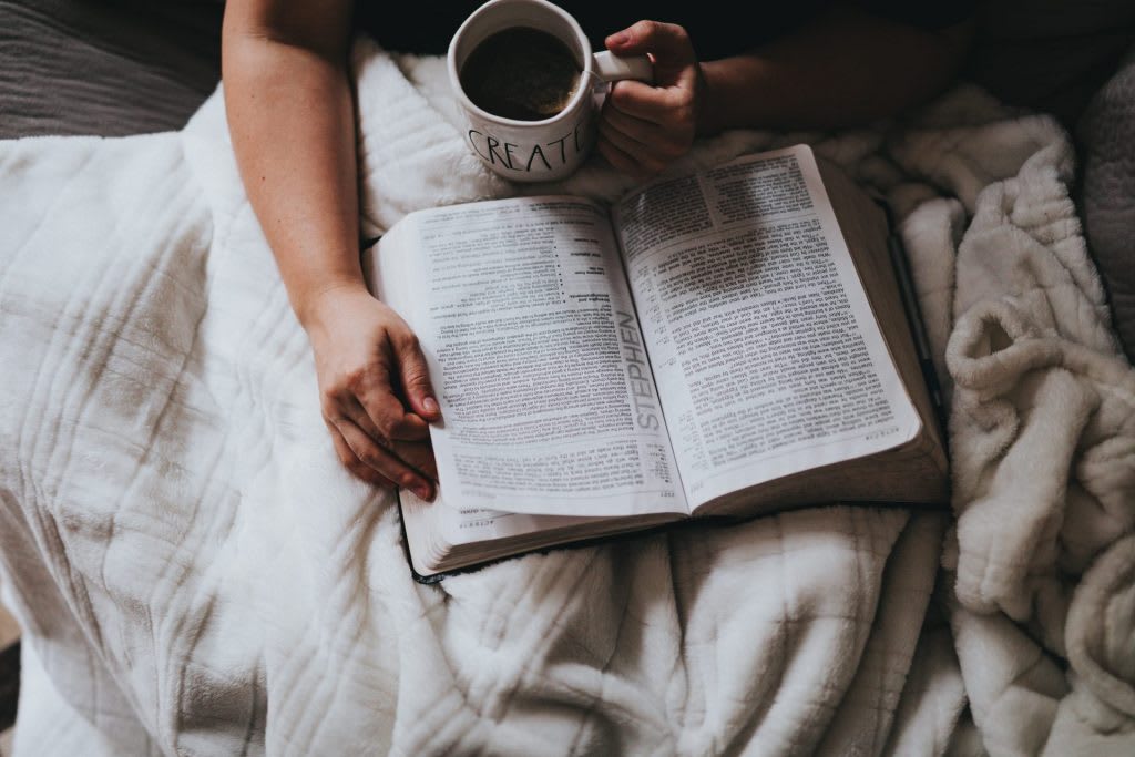Some sits on a couch covered in a blanket, holding a cup of tea and a bible.
