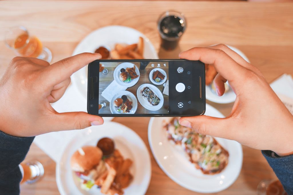 Two hands holding an iPhone over a table of food, taking a picture of it.