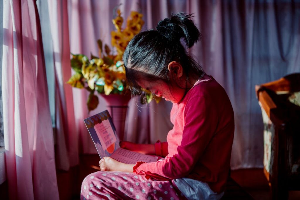 Karunia sits in her room reading a letter. Her windows are surrounded in pink curtains.