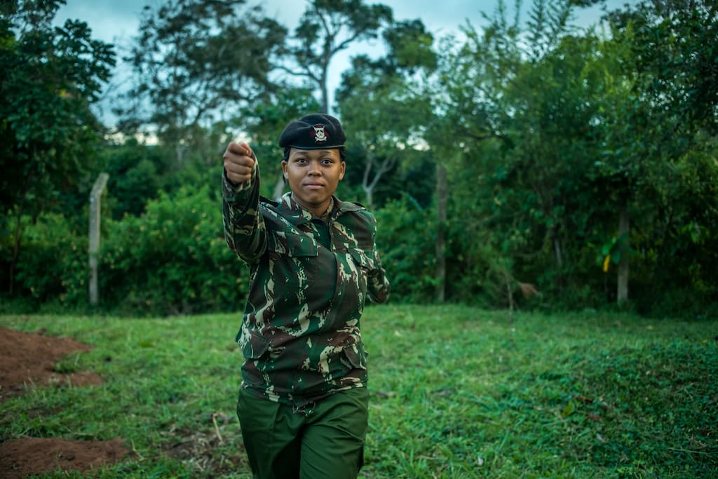 Miriam is standing in a field in front of trees. She is wearing a camoflauge shirt and a black hat. Miriam is looking at the camera and has her arm outstretched.