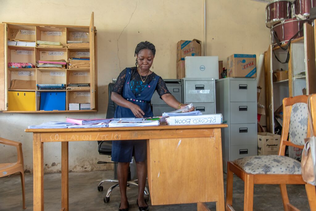 This is Emmanuella, a collation officer for the Tongu cluster. She receives all the cluster’s letters and distributes them when the projects representatives come. She is standing behind a desk and is wearing a blue dress. There are filing cabinets in the background.