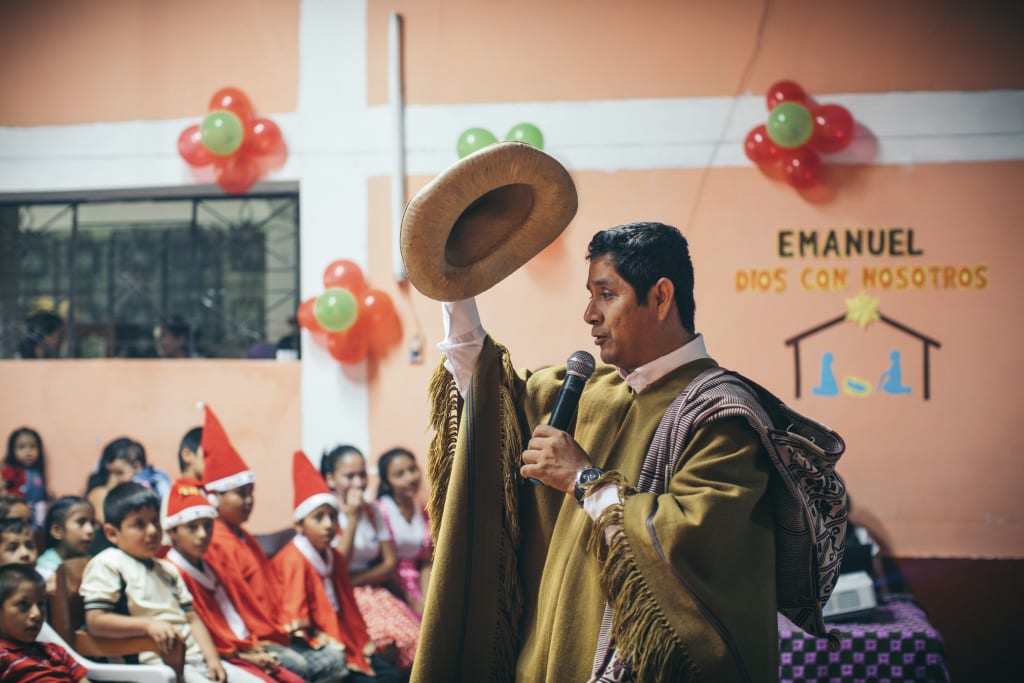 A pastor wears a traditional outfit, tipping his hat as he shares a Christmas message with the children.