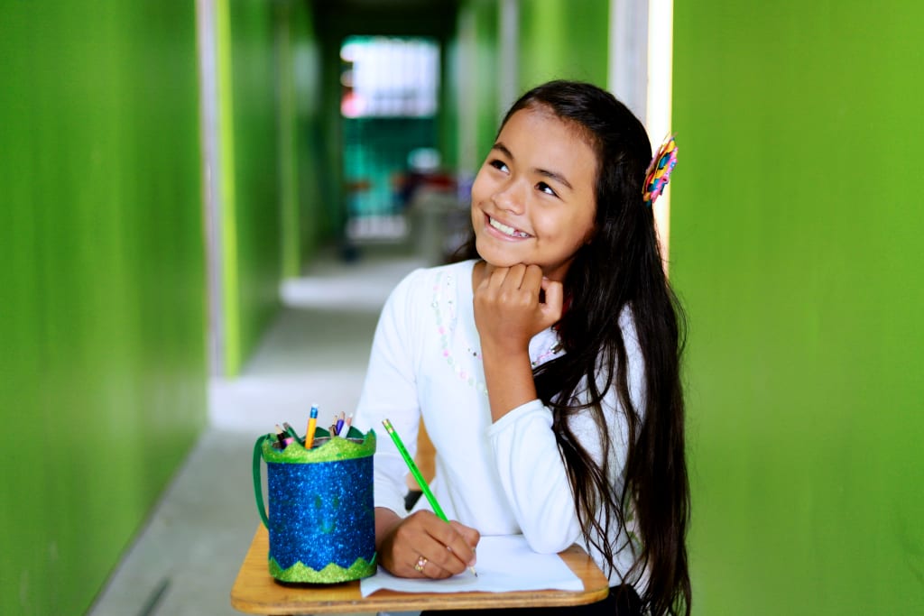 A young girl, Nazareth, sits at a desk with her chin on her first, looking up as she thinks. She is holding a green pencil.