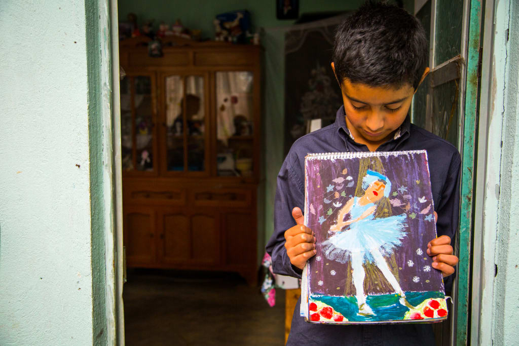 Hector, a young boy, stands in a doorway, looking down at his painting of a ballerina, which he holds up to his chest.