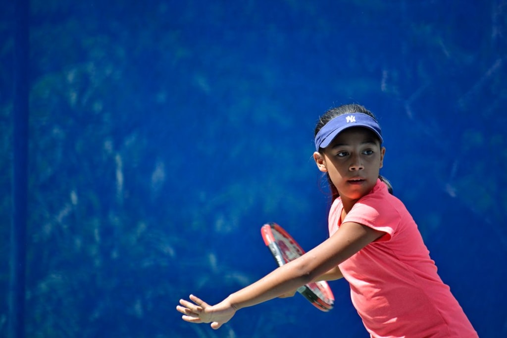 Alondra, wearing a pink shirt and blue visor, winds up with her tennis racquet to hit a ball.
