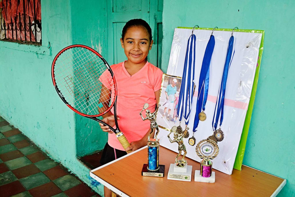 Alondra, wearing a pink shirt and holding her tennis racquet, smiles and poses with her medals and trophies.