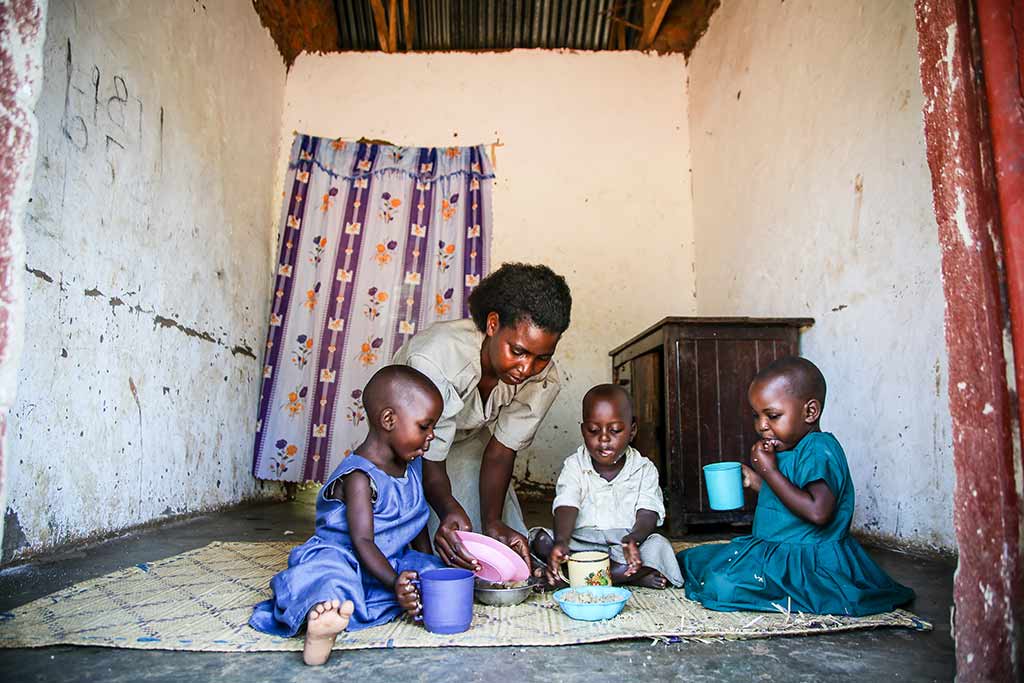Three children sit on a mat inside a small room eating and drinking, while their mom helps.