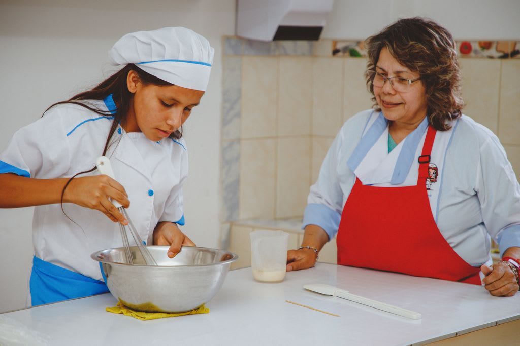 A girl in a white coat and white baker's cap uses a whisk in a metal bowl while a woman in a red apron watches.