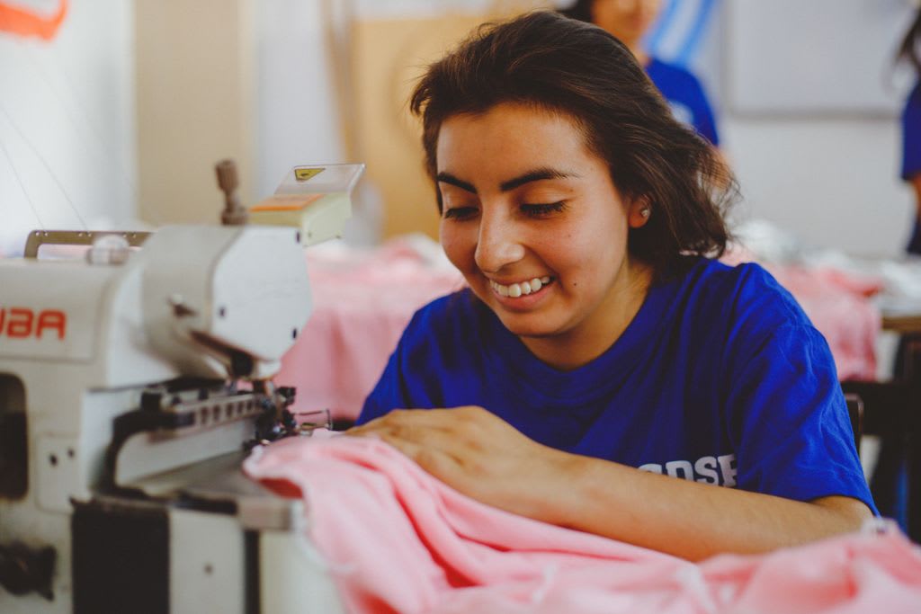 A girl wearing a blue shirt smiles as she sews pink cloth on a sewing machine.
