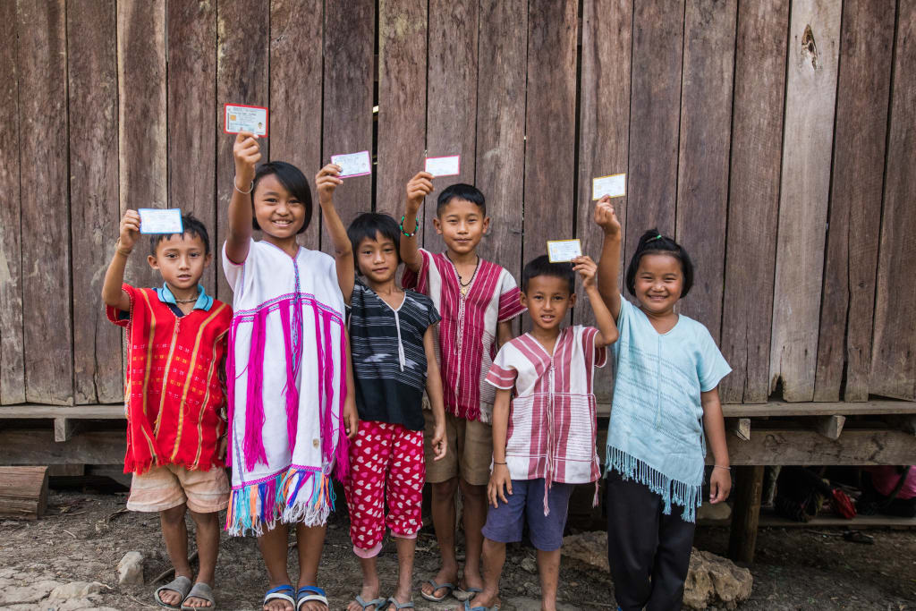 Six children smiling against a fence holding their Thai citizen cards.