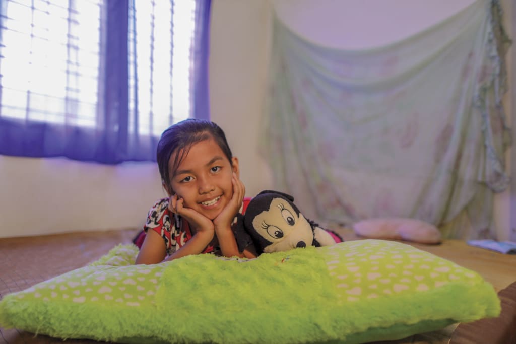 8- year-old girl lies on her pillow with a stuffed animal, smiling and posing for the camera.