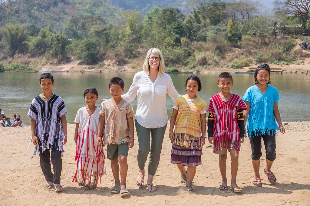A woman in white stands in the middle of a group of children wearing colorful tribal clothing in front of a river and mountains.