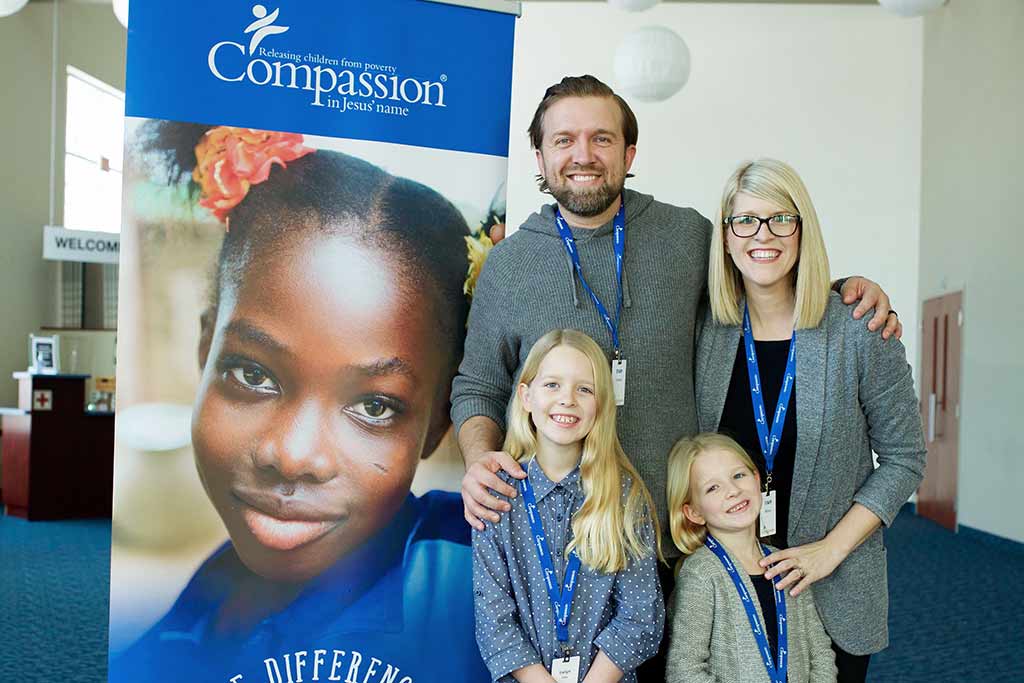 A smiling family of four stands in front of a poster that says Compassion.